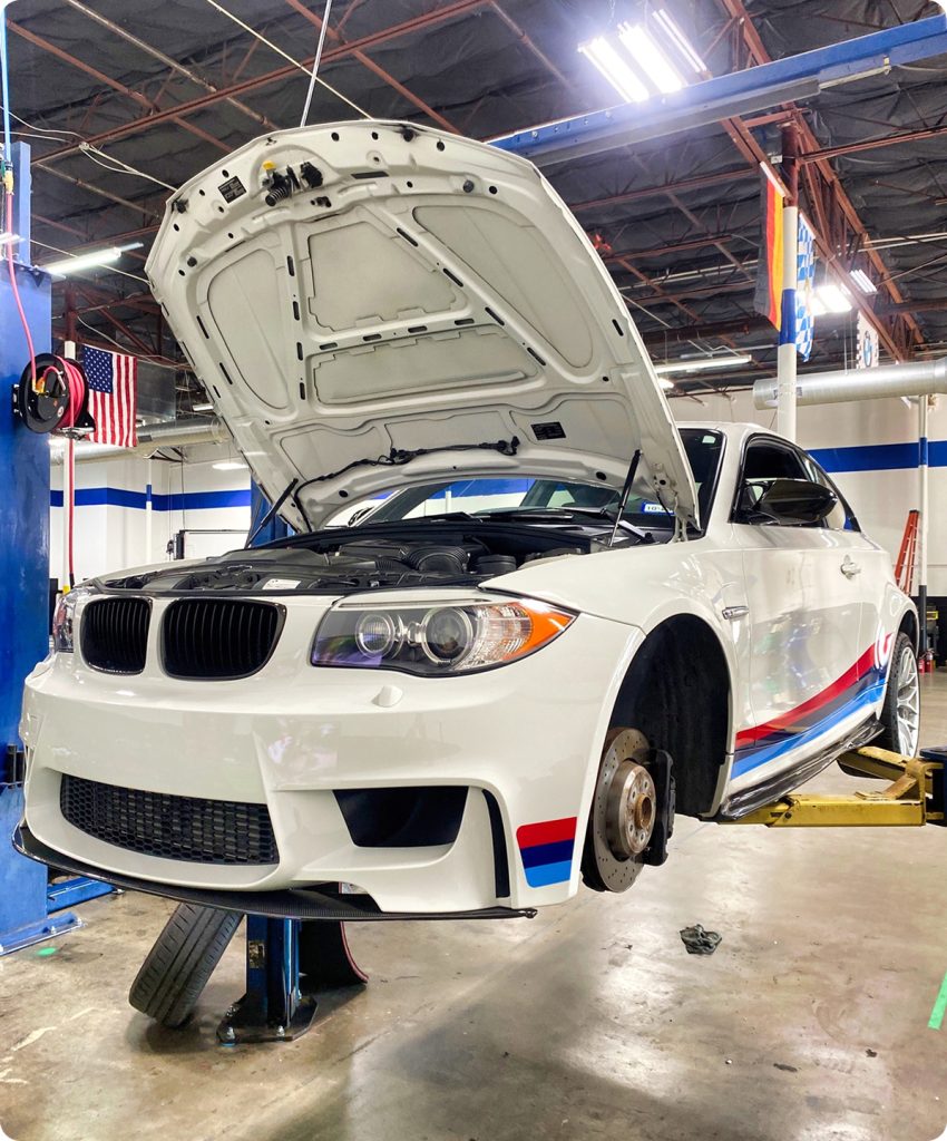 White BMW 1 Series M Coupe with M-performance livery on a hydraulic lift for brake service and engine maintenance at Ultimate Bimmer Service.